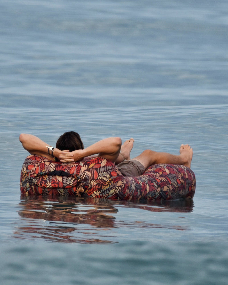 A person relaxes on their back inside a patterned red, orange, and black inner tube float on calm blue water.